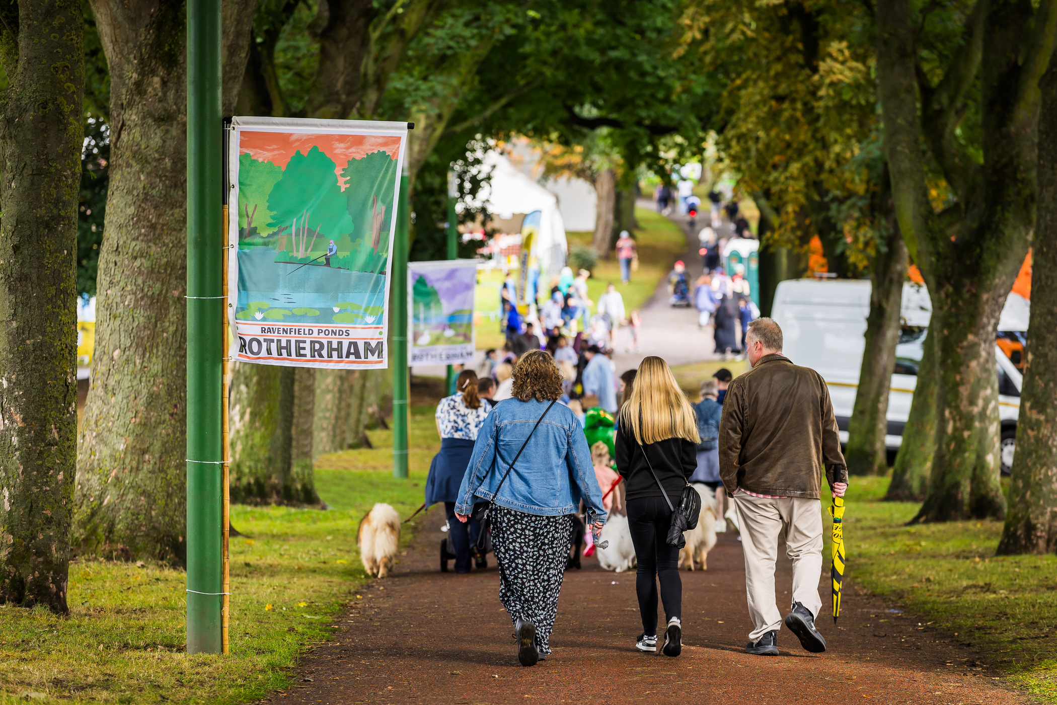 A photo of people walking through Clifton Park during Rotherham Show. There is a flag hung to a pole and a large tarmac path.